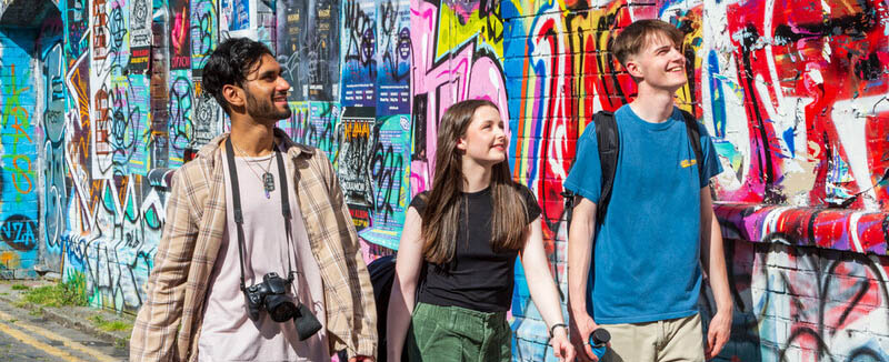 three students walk down the pavement in stokes croft, with a graffiti decorated wall alongside them